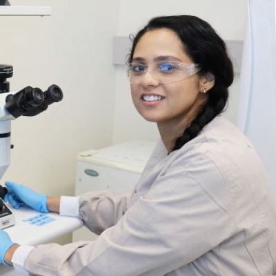 a woman with dark hair wearing safety glasses sits in front of a white and black microscope